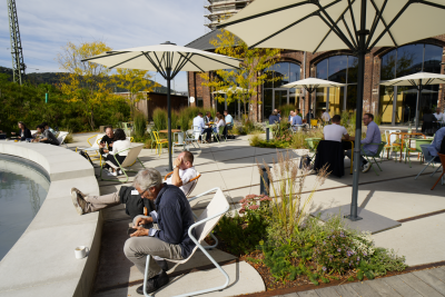 Mittagspause beim Mobilitätskongress. Auf der Terrasse des Lokschuppens in Marburg sitzen die Teilnehmer an Tischen und essen. Es sind große weiße Schirme aufgespannt. Links eine Wasserfläche. Vier Personen sitzen an der Wasserfläche und schauen auf ihre Smartphones oder telefonieren. Im Hintergrund ein Backsteingebäude mit großen Fenstern.