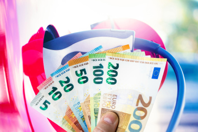 Eine Hand h&auml;lt einige Geldscheine vor einem Ladestecker, der in einem eAuto steckt.