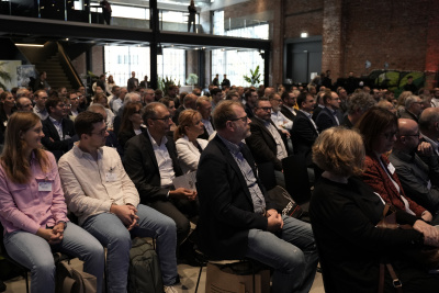 Eine große Gruppe Menschen sitzen in einem Saal auf Stühlen in Reihenbestuhlung und schauen nach vorne