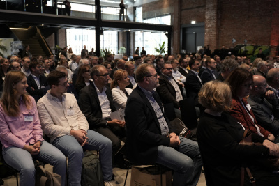 Eine große Gruppe Menschen sitzt in einem Saal auf Stühlen in Reihenbestuhlung und schauen nach vorne Richtung Bühne