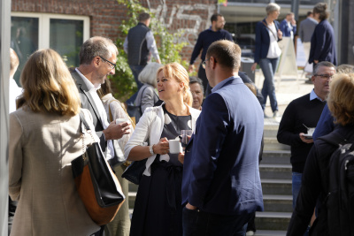 Pause beim Mobilitätskongress. Im Vordergrund steht eine Gruppe aus zwei Männern und eine Frau. Sie stehen draußen vor dem Gebäude und unterhalten sich. Sie haben jeweils eine Kaffeetasse in der Hand. Links, rechts und im Hintergrund sind weitere Personen die umherlaufen oder sich unterhalten und Kaffee trinken.