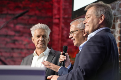 3 Männer stehen auf einer Bühne, einer spricht ins Mikrofon, Blick ins Publikum