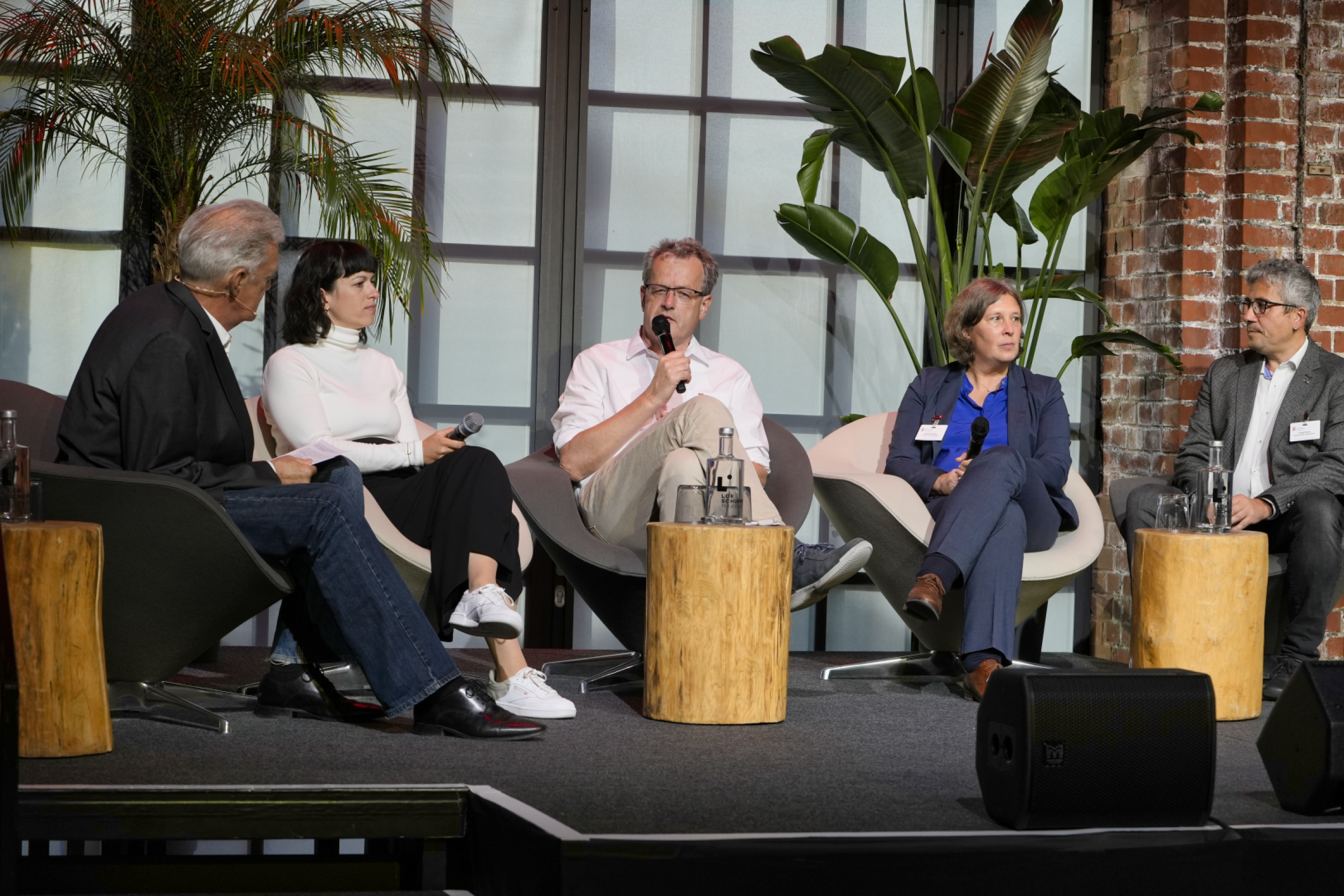 Podiumsdiskussion auf der Bühne im Lokschuppen Marburg. Es sitzen zwei Frauen und drei Männer auf Sesseln mit kleinen Stehtischen auf denen Getränke stehen. Ein Mann spricht in ein Mikrofon, die anderen hören zu. Im Hintergrund stehen Pflanzen zur Dekoration.