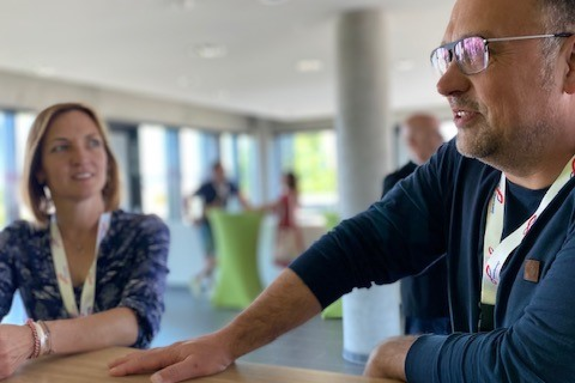 Das Bild zeigt einen Mann im Vordergrund an einem Tisch auf dem ein Klemmbrett mit Unterlagen liegt. Er spricht. Im Hintergrund eine Frau die den Mann anschaut.