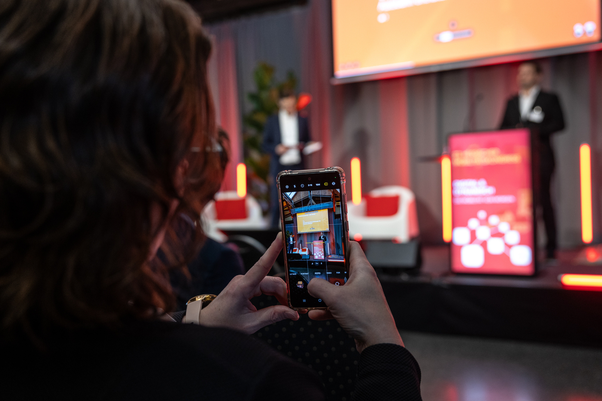 Foto 7 Mobilitätskongress 2025 Teilnehmer macht ein Handyfoto, Bühne mit Referent im Hintergrund