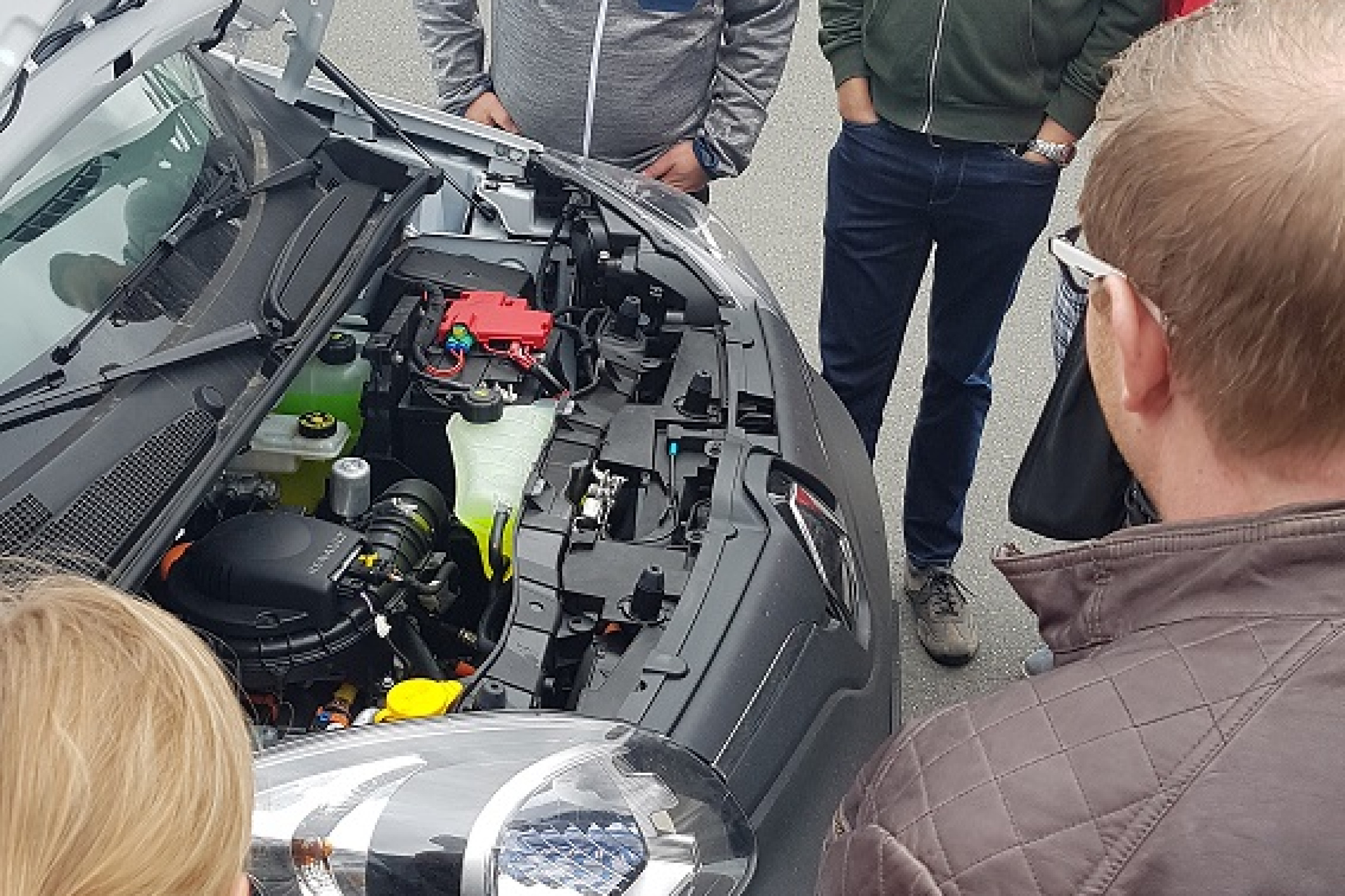 Sechs Teilnehmer stehen auf einem Parkplatz um einen Elektroauto und schauen in die geöffnete Motorhaube. Im Hintergrund parken zwei Müllsammelfahrzeuge.