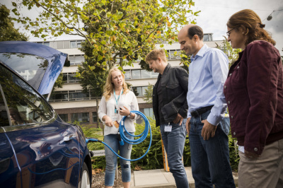 Seminarleiterin erklärt das Laden eines E-Autos. Sie hat ein Ladekabel aufgewickelt, der Ladestecker steckt in der Ladebuchse des Elektroautos. Drei weiterer Personen stehen am Auto und hören der Seminarleiterin zu.